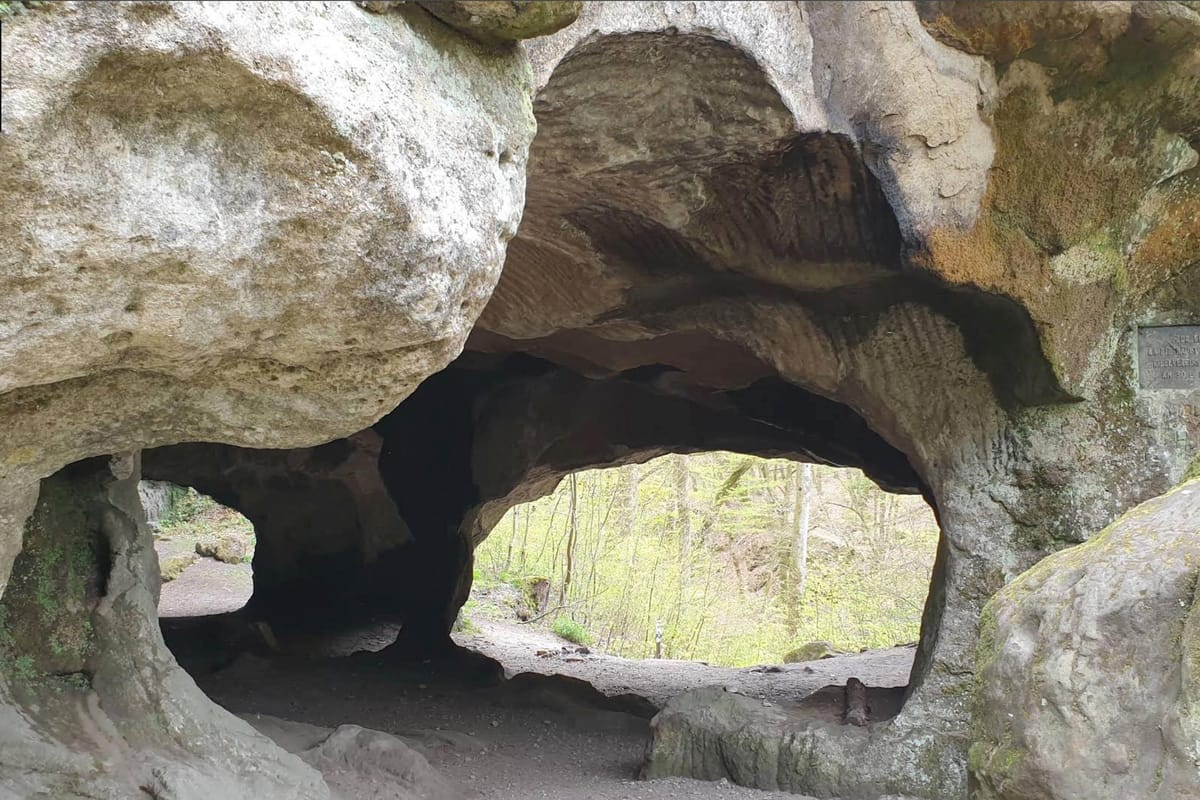 Natural sandstone rock formation with cave-like arches framing a green forest hiking trail near Berdorf